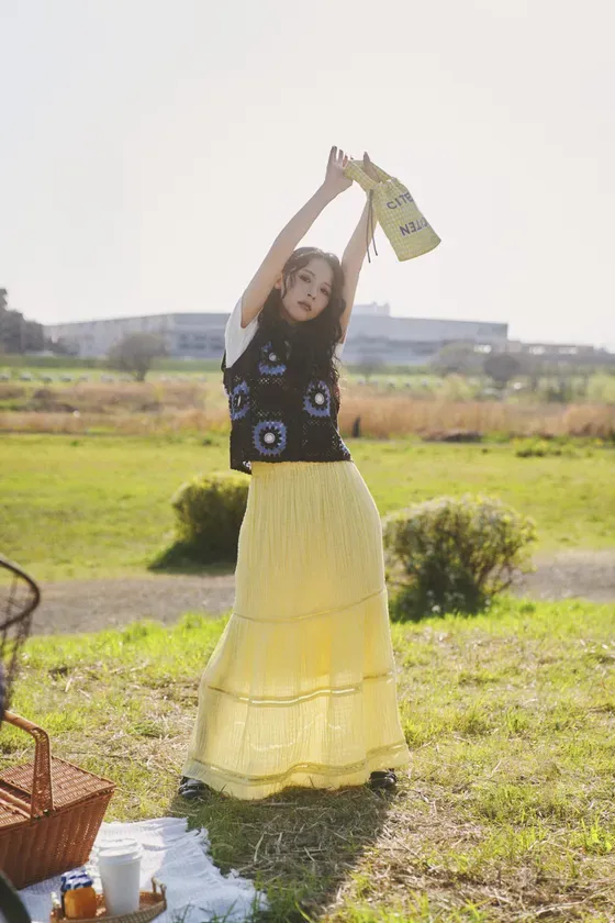 女性が野原で両手を上げてバッグを持っている写真。彼女は黒のベスト、黄色のスカート、白いシャツを着ています。ピクニックバスケットもあります。