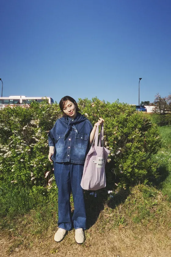 青空の下、デニムのセットアップにチェック柄のトートバッグを持った女性が立っている様子。