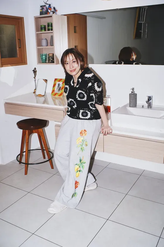 洗面台に寄り添う女性の写真。彼女は黒と白のセーターと花の刺繍が入ったグレーのズボンを着用している。背景には鏡、棚、洗面台、スツールなどがある。