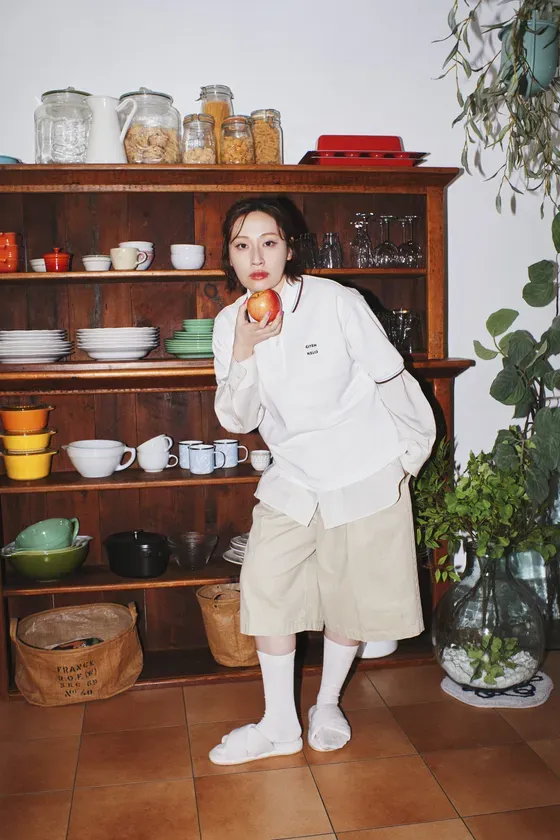 女性がリンゴを持ち、キッチンに立っている写真。木製の食器棚には、食器、食材、グラスなどが置かれている。女性は白いシャツとベージュのショートパンツを着ており、足元はサンダル。背景には観葉植物がある。