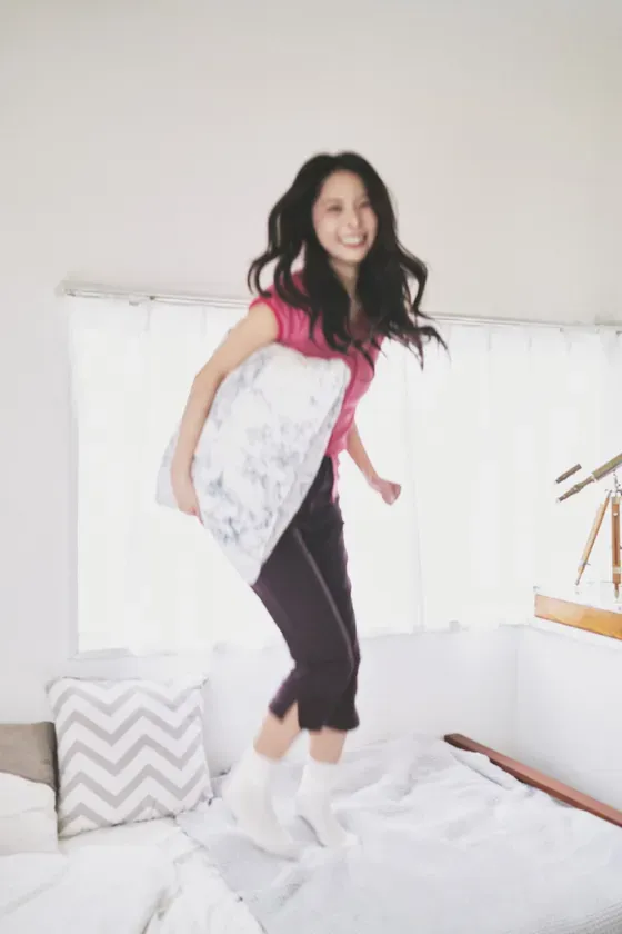 ベッドの上でジャンプする女性の写真です。彼女はピンクのシャツと黒いズボン、白い靴下を履いています。彼女は枕を抱えています。背景には白いカーテンのある窓と望遠鏡があります。