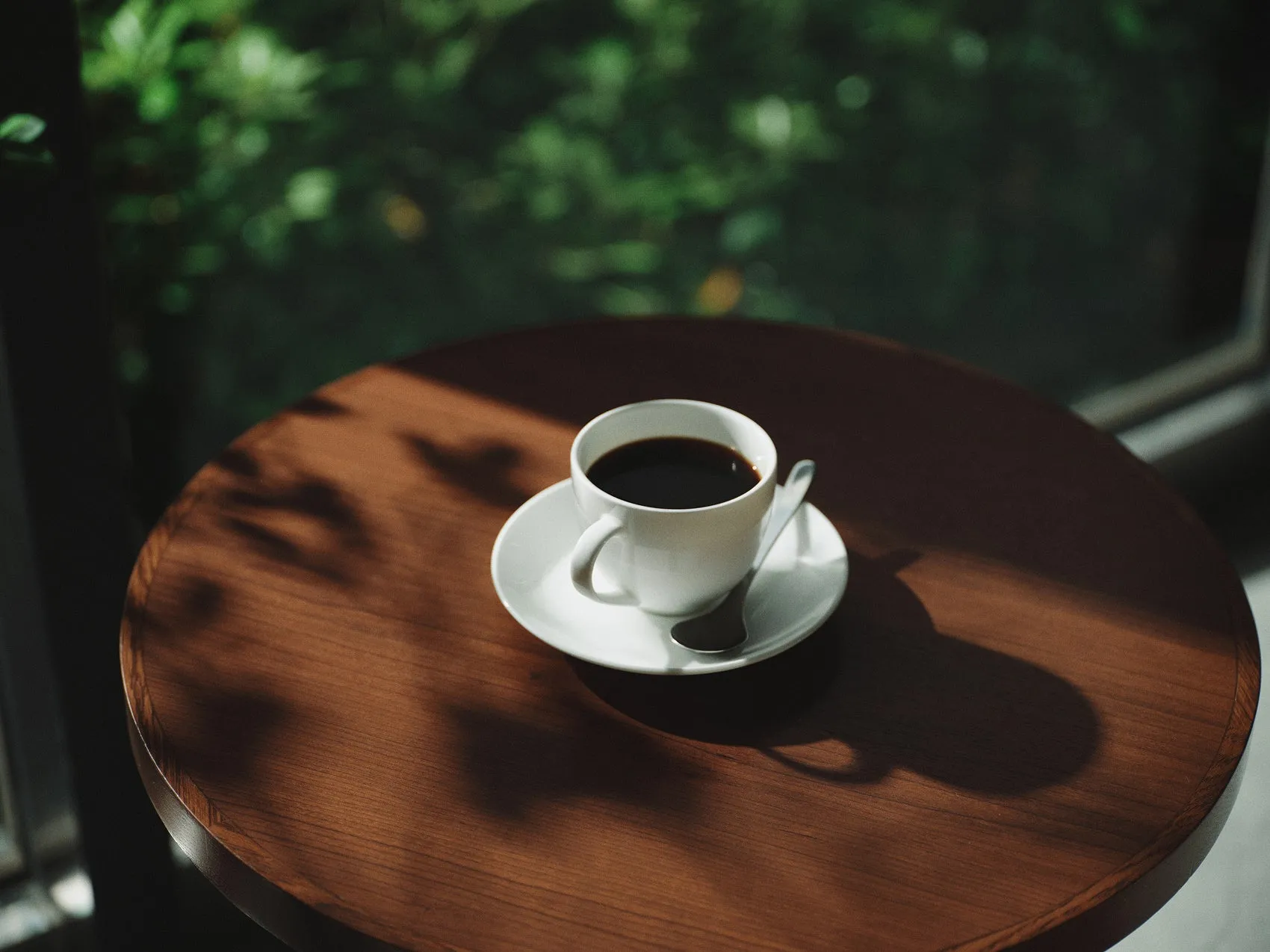 木製のテーブルに置かれたコーヒーカップの写真。カップは白く、コーヒーが注がれている。背景にはぼやけた緑の木々が見える。
