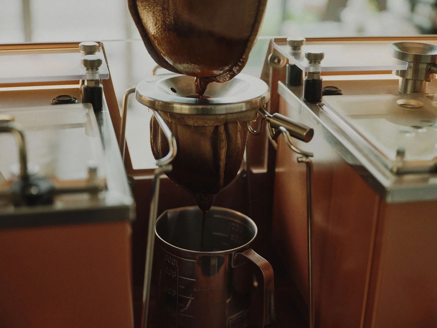 コーヒーを抽出している様子を捉えた写真です。コーヒーメーカーからコーヒーが抽出され、金属製のカップに落ちています。コーヒーメーカーとカップは、木製のフレームに囲まれた、透明なガラス面の上に置かれています。