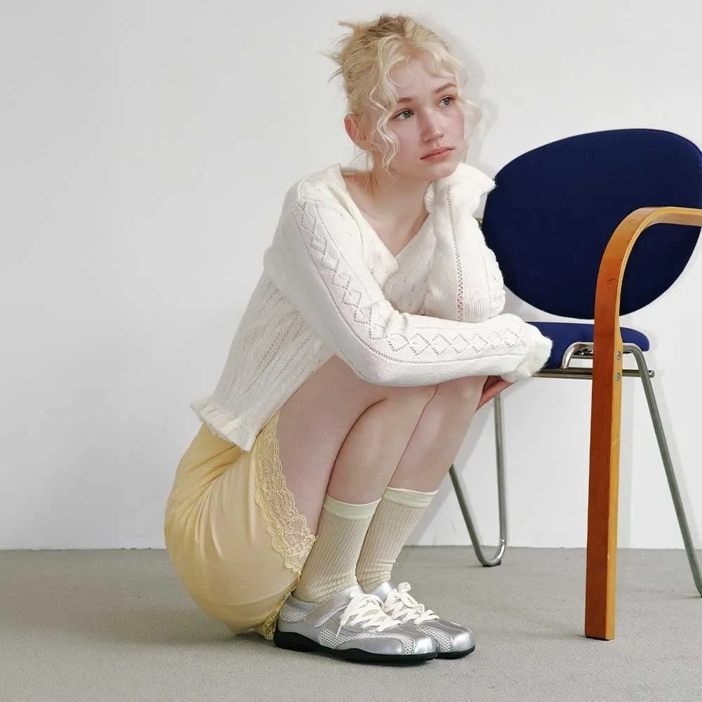 若い女性がしゃがみこんでいる写真。彼女は白いセーター、黄色のスカート、靴下、スニーカーを履いている。背景には青い椅子と白い壁がある。