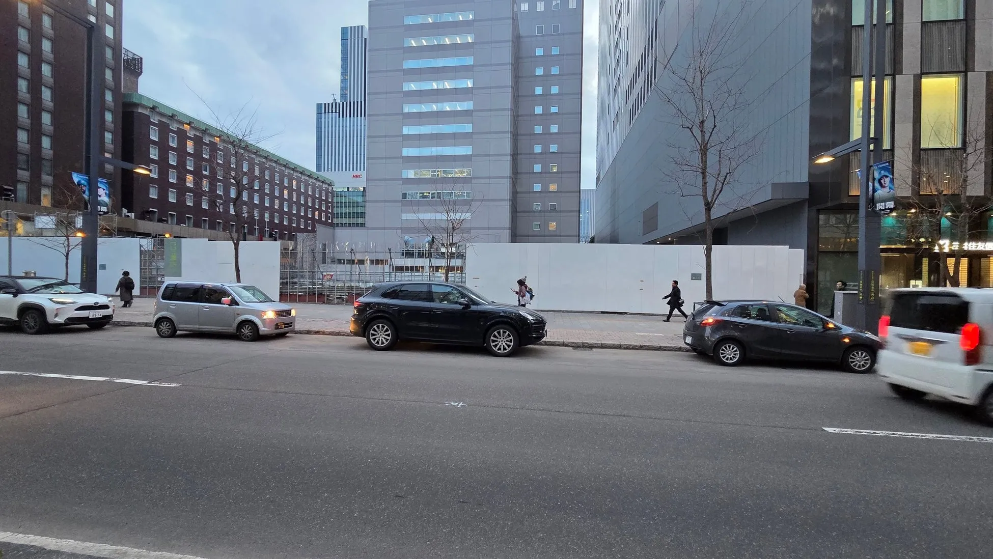 街並みを写した写真。道路に面して車が複数台駐車されており、歩道を人が歩いています。背景には高層ビルがそびえ立っています。