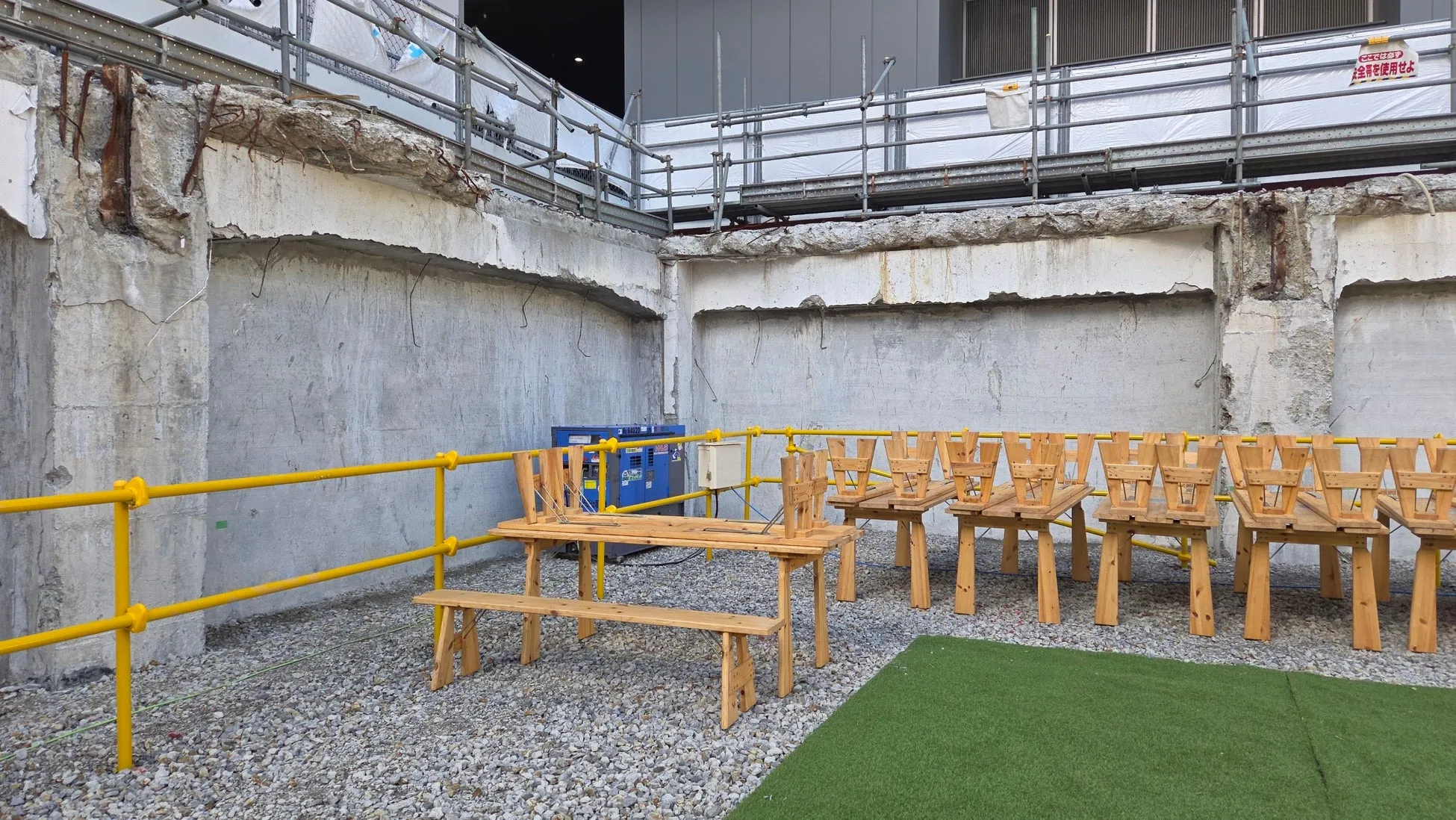 建設現場と思われる場所で、木製のテーブルと椅子が置かれています。壁はコンクリートで覆われ、足場が組まれています。