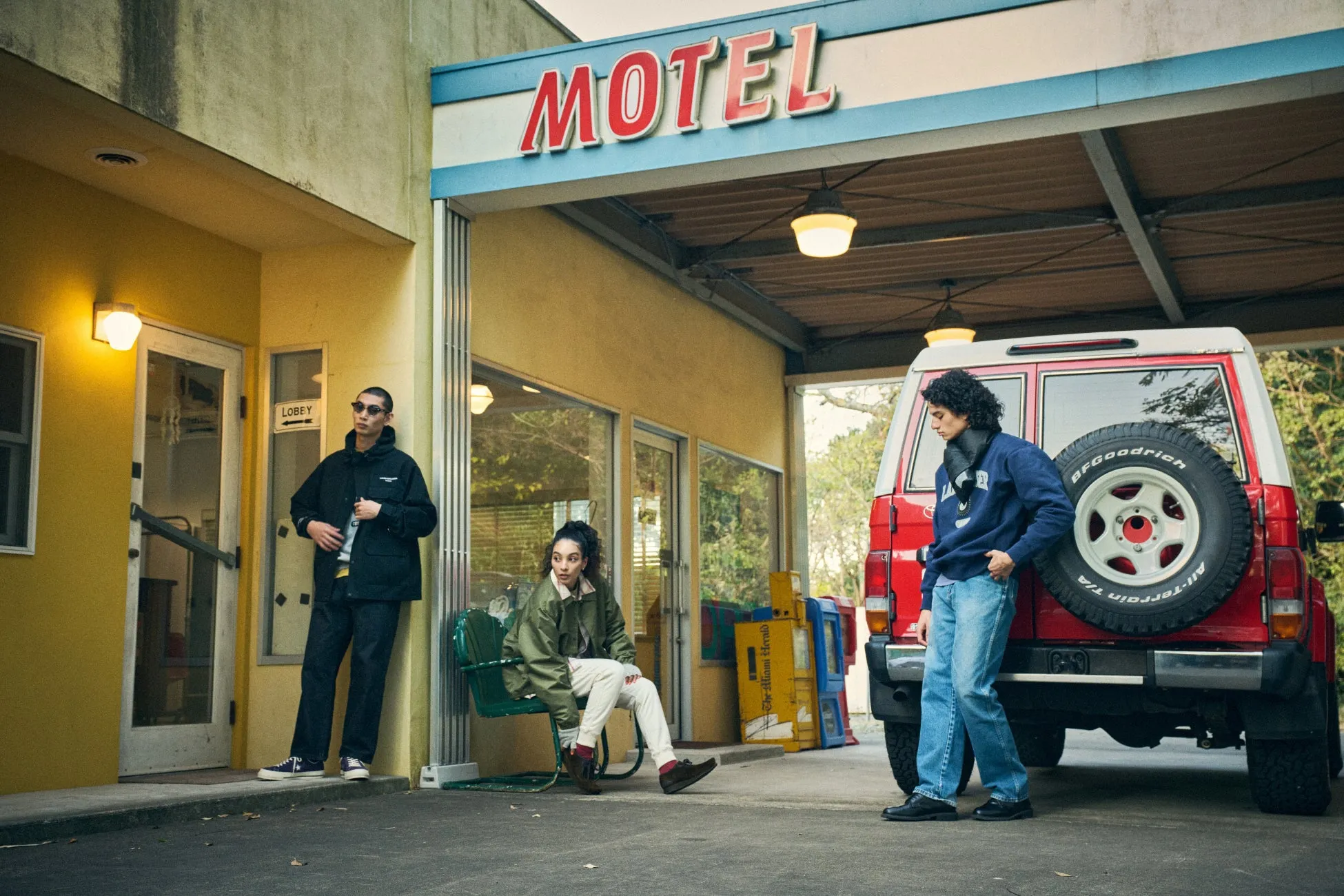 モーテルの前に立つ3人の人物。赤い車が駐車場に止まっている。