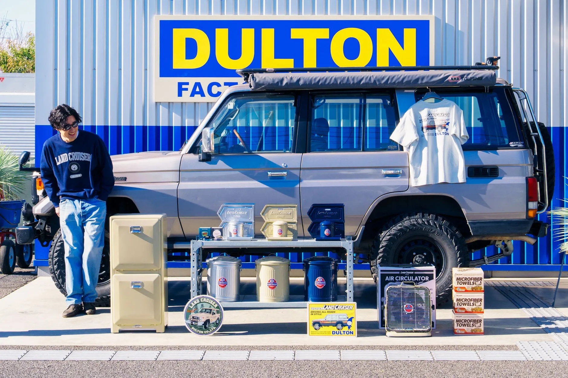 DULTONのロゴが入った建物とランドクルーザーの車、関連グッズを並べた写真。男性が写っており、車やグッズを眺めている様子。
