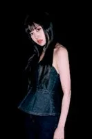 黒い背景に、デニムのトップスとジーンズを着た女性のポートレート。女性はカメラを見ており、髪は黒く、前髪がある。