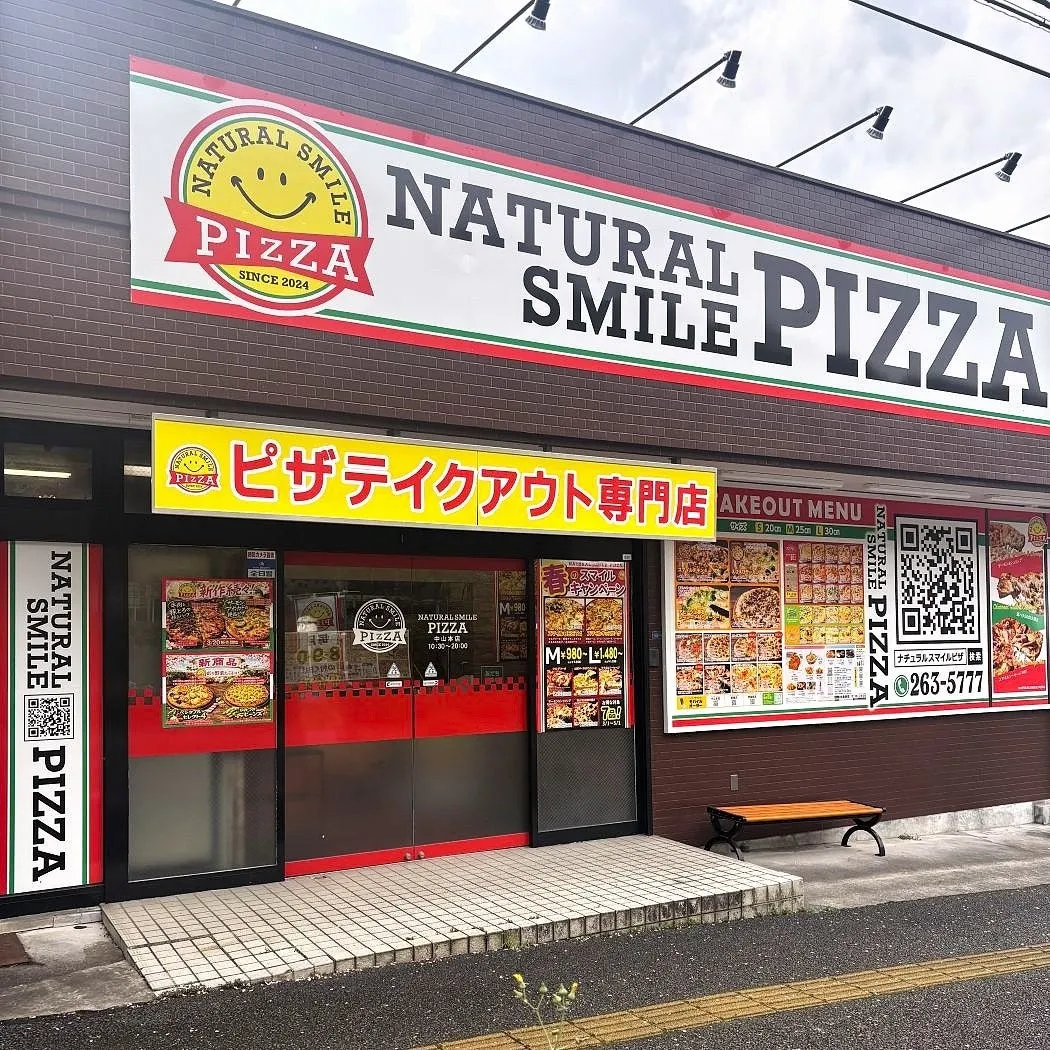 ピザテイクアウト専門店「NATURAL SMILE PIZZA」の外観写真。看板には「NATURAL SMILE PIZZA」のロゴと「SINCE 2024」の文字、そして「ピザテイクアウト専門店」の文字が記されています。店内にはメニューやQRコードが表示されています。