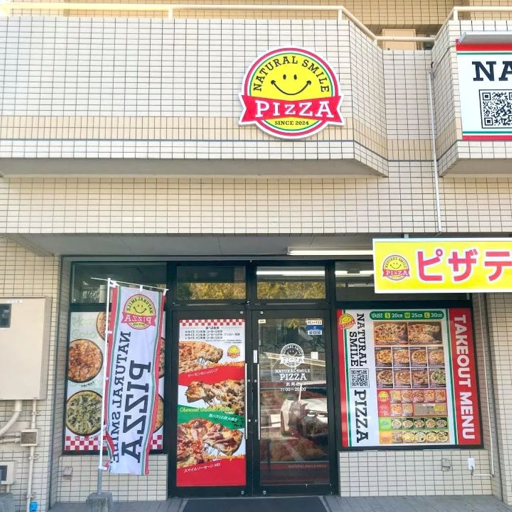ピザ屋の正面玄関の写真。看板には「NATURAL SMILE PIZZA」と書かれており、テイクアウトメニューも表示されている。