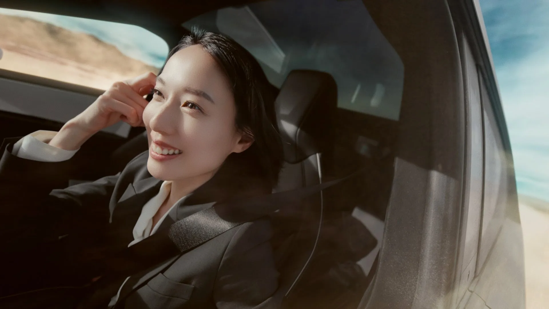 車の中から笑顔で外を眺める女性。ジャケットを着ており、髪は黒髪で、日差しを浴びている。