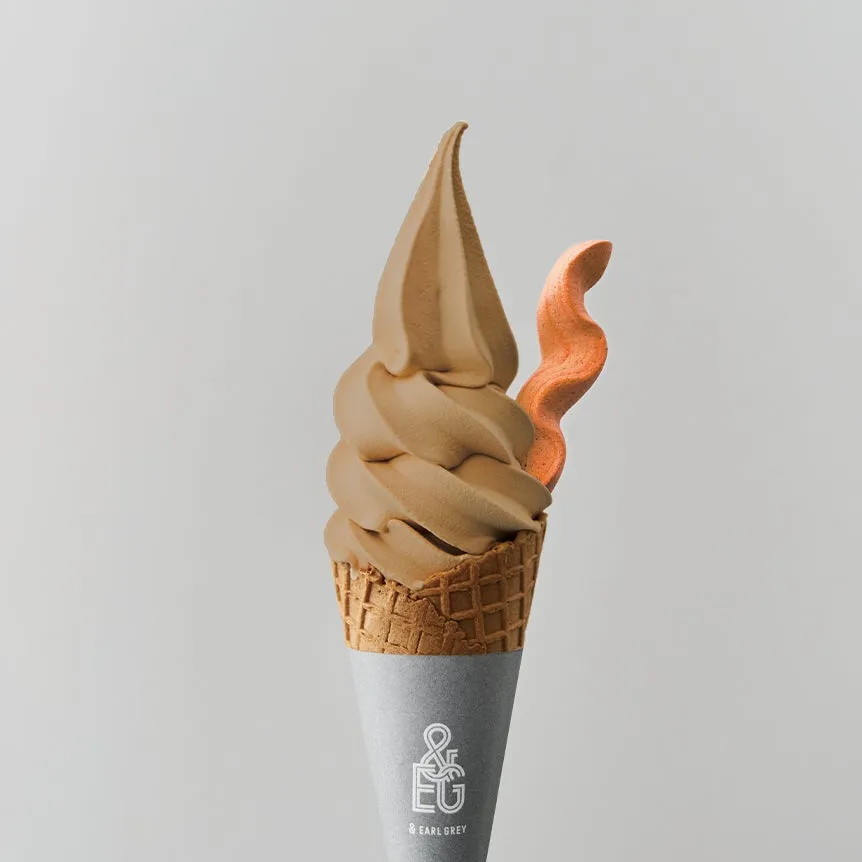 ソフトクリームのクローズアップ。ソフトクリームは茶色で、コーンにオレンジ色の装飾が添えられています。背景は白です。