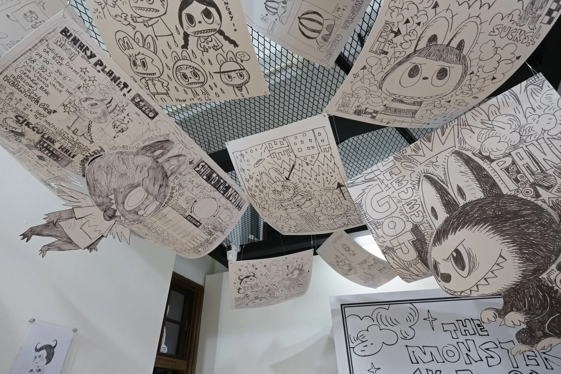 展示されている、手描きのイラストが描かれた複数の紙。それぞれの紙には、キャラクター、幾何学模様、テキスト、その他のデザインが描かれています。