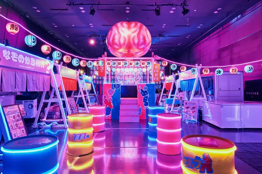 ネオンカラーが印象的な、日本の屋台のような雰囲気のイベント会場の写真。中央には大きなオブジェがあり、提灯や屋台、カラフルなライトで装飾されている。
