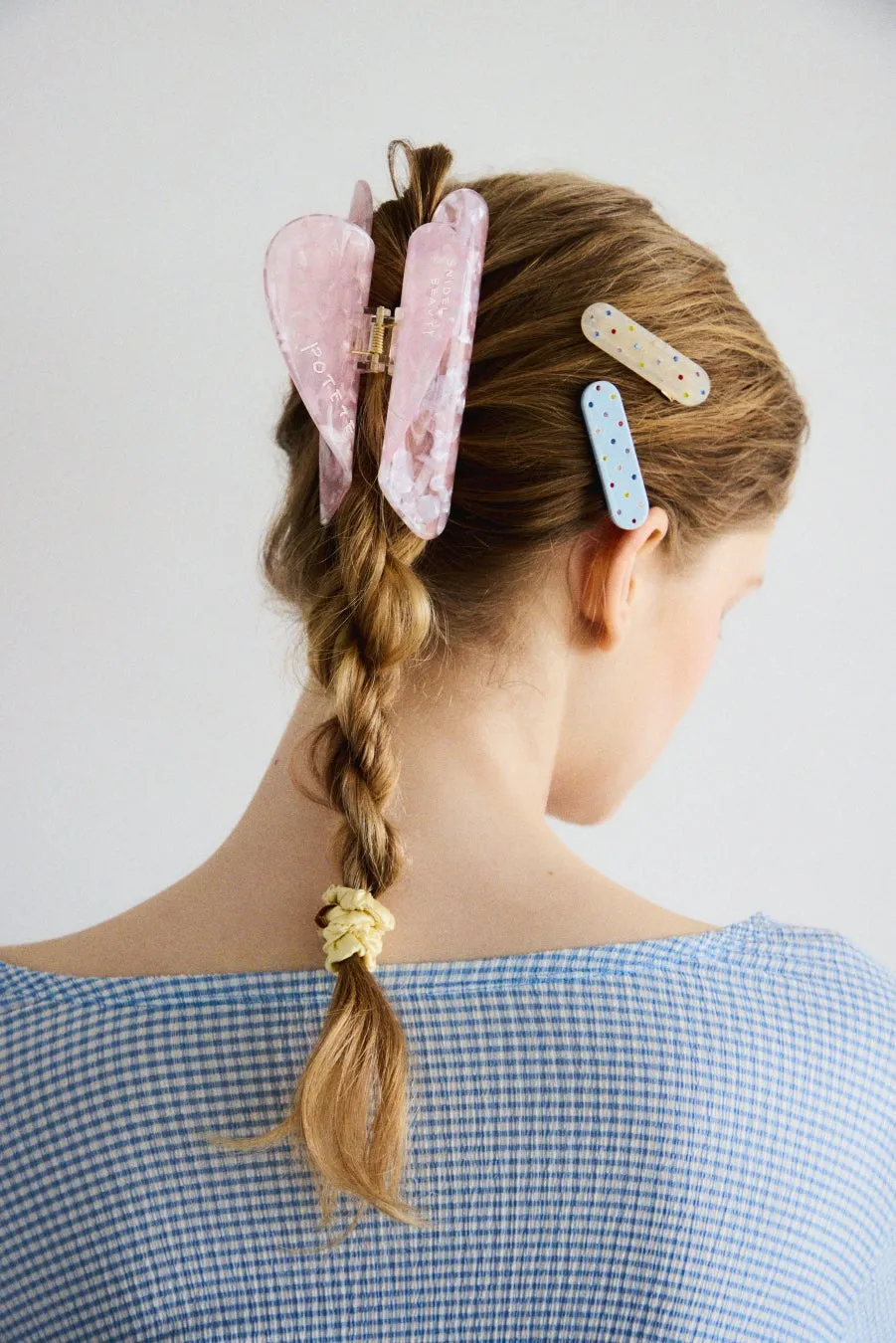 後ろ姿の女性。髪は三つ編みで、ヘアクリップやヘアピンで飾られている。青と白のチェック柄の服を着ている。