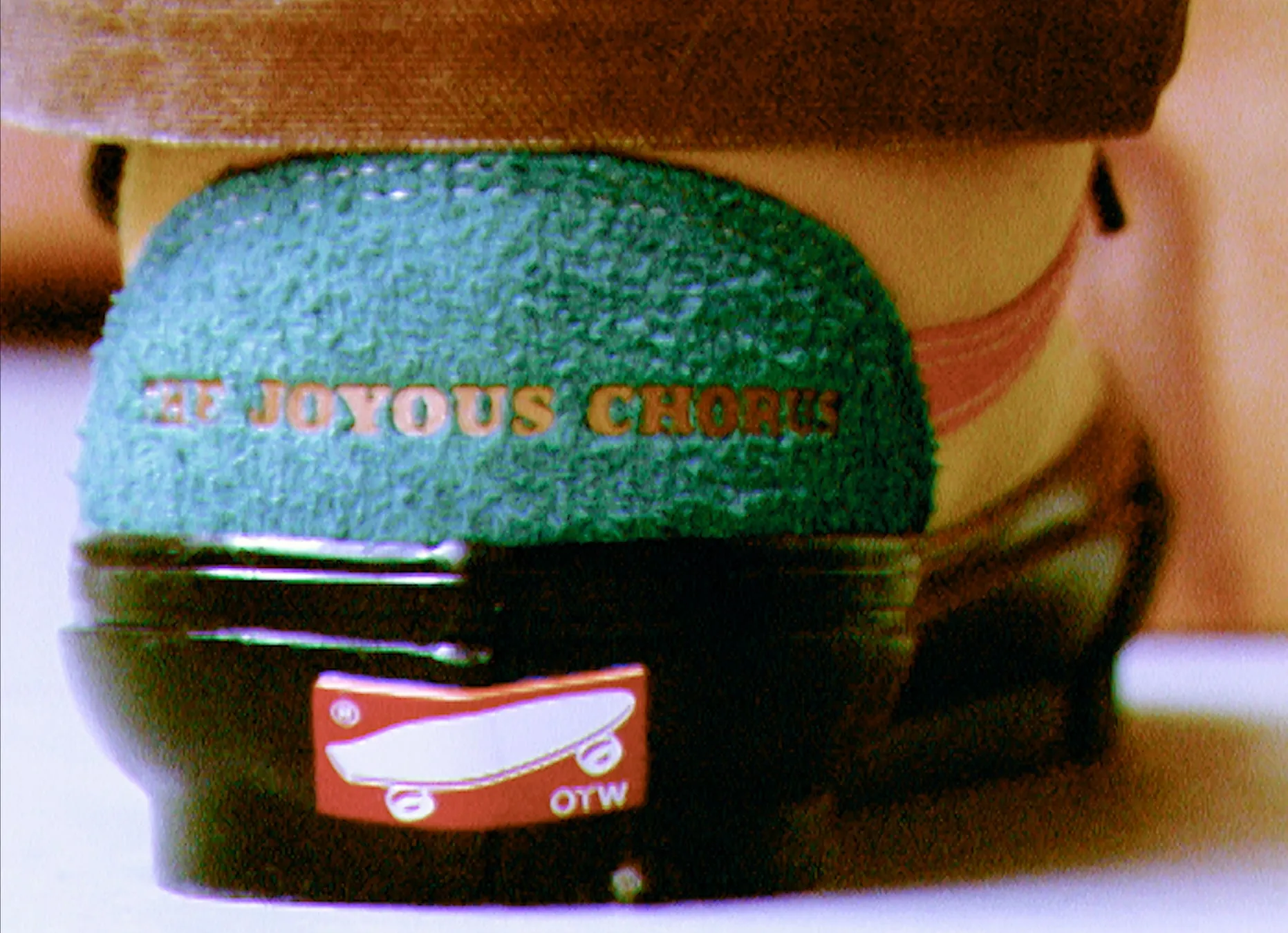 靴の後部にあるロゴと文字が写っているクローズアップ画像です。緑色の部分に「THE JOYOUS CHORUS」と書かれており、その下の黒い部分には「OTW」のロゴがあります。