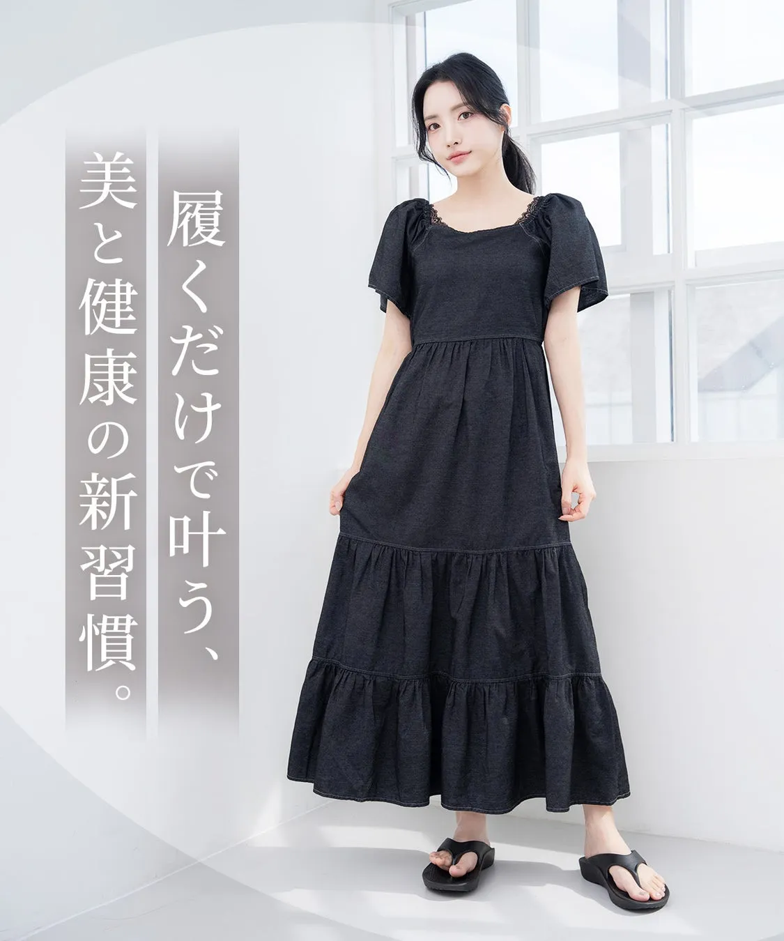 窓際に立つ女性が、黒いワンピースを着て立っている写真です。ワンピースは、パフスリーブで、スカート部分はティアードデザインです。女性は黒いサンダルを履いています。