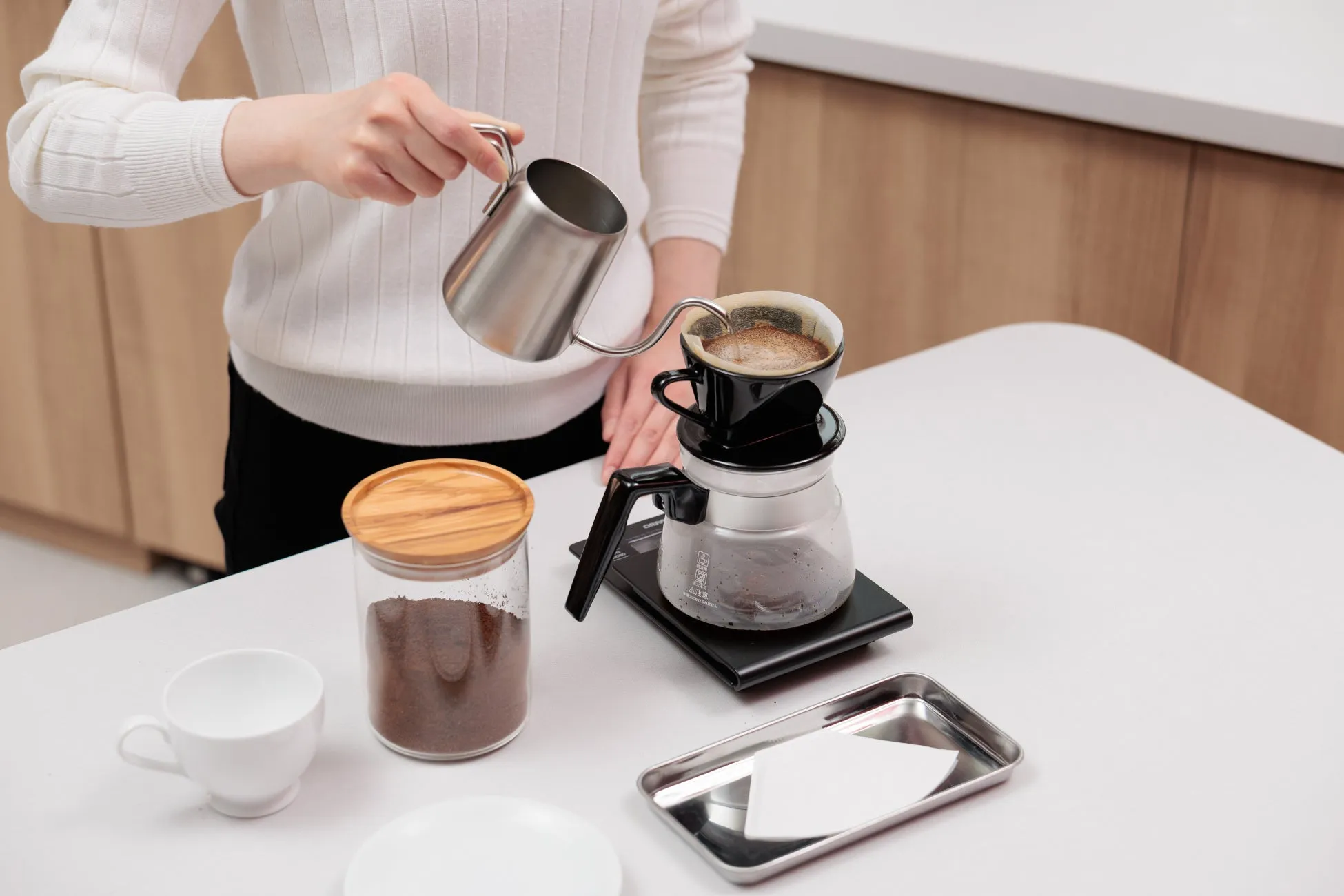 コーヒーを淹れている様子。女性がケトルからドリッパーに湯を注いでいる。コーヒー豆、カップ、サーバー、ペーパーフィルターなど、コーヒーを淹れるための道具が写っている。