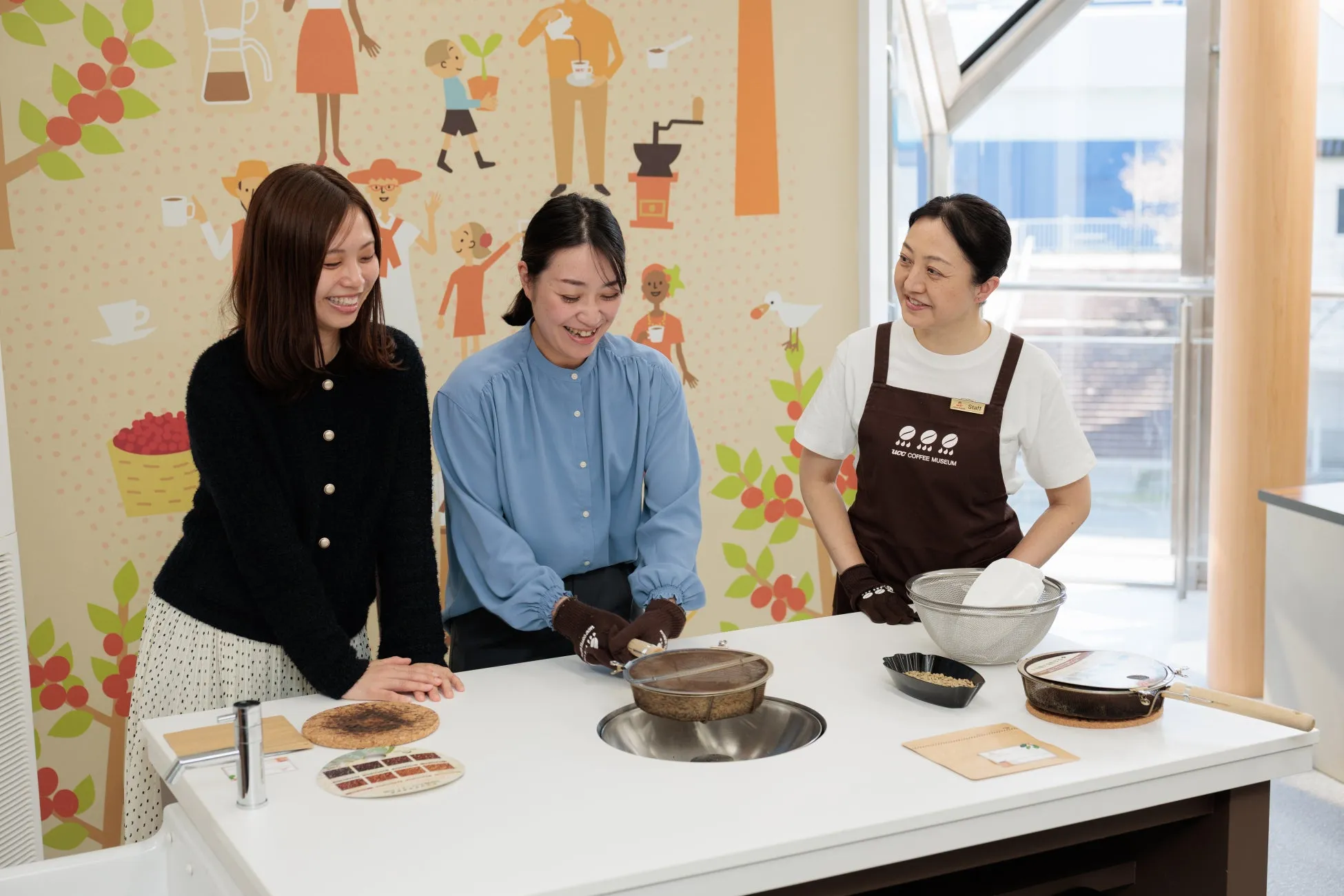 コーヒー教室の様子。3人の女性がテーブルを囲み、コーヒー豆の焙煎作業をしている。壁にはコーヒーに関するイラストが描かれている。