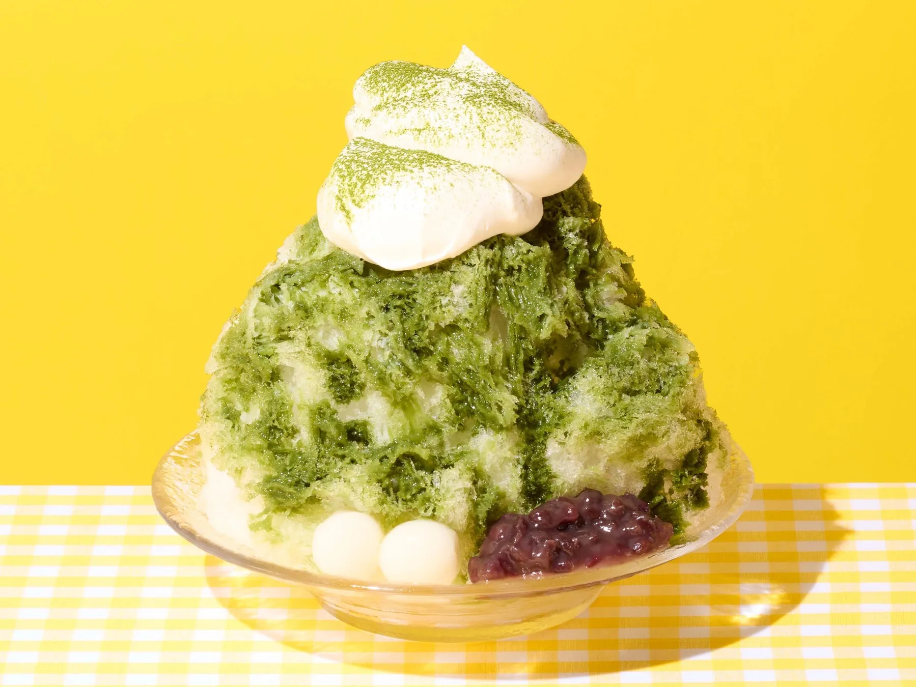 抹茶のかき氷の写真。白いクリームがトッピングされ、横に白玉とあんこが添えられている。背景は黄色で、テーブルクロスは黄色のチェック柄。