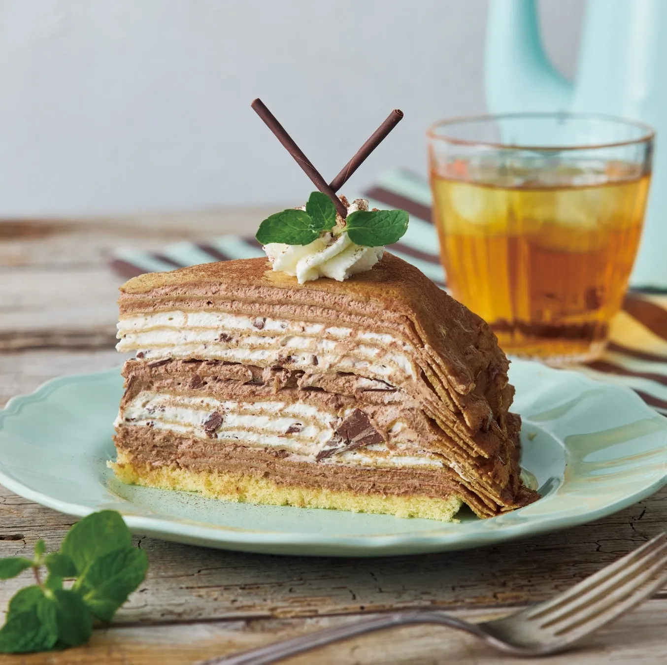チョコレートクレープケーキと飲み物の写真。ケーキはミントの葉で飾られ、チョコレートのスティックが添えられています。テーブルの上にはフォークも置かれています。