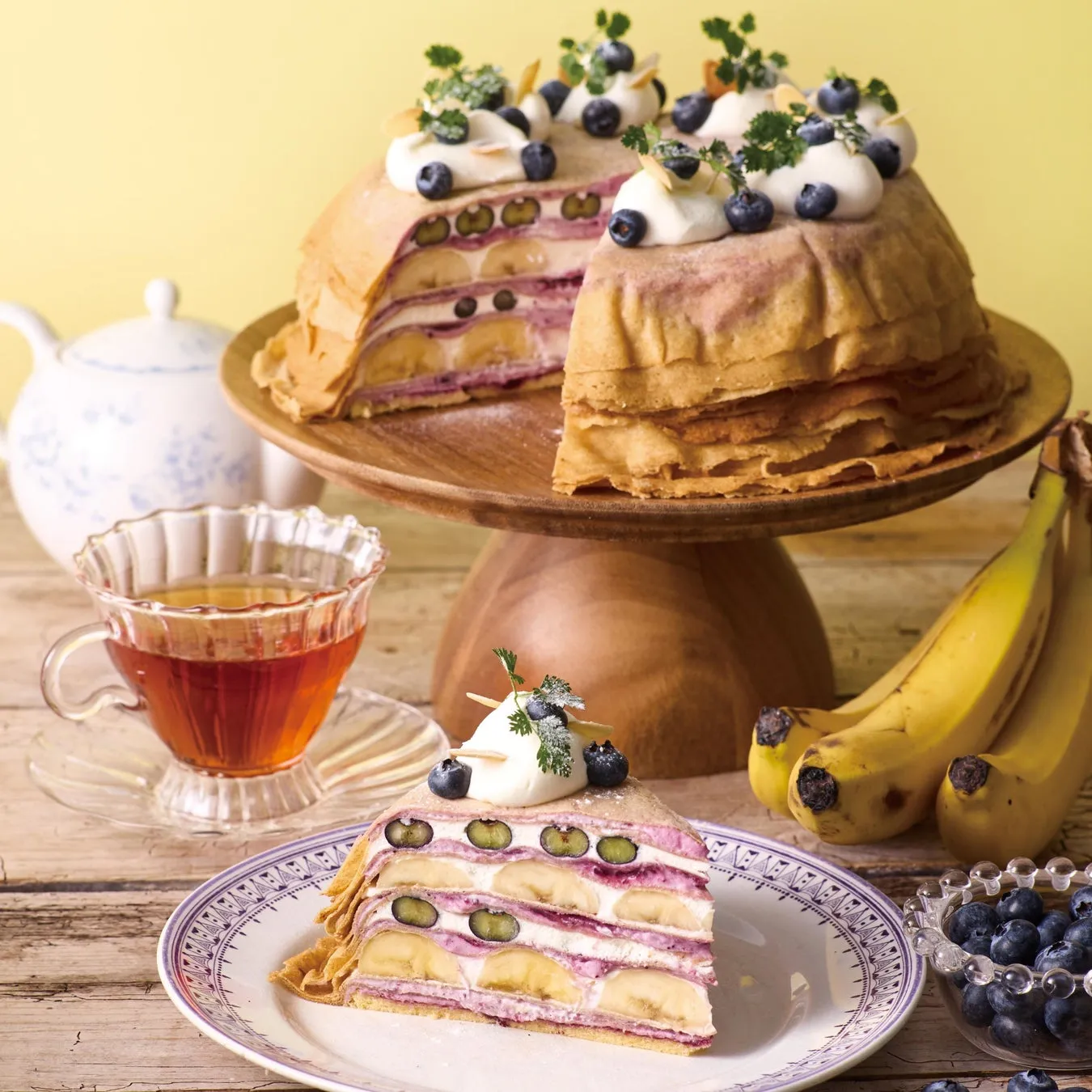 ブルーベリーとバナナが挟まれたクレープケーキがメインの画像です。ケーキの横には紅茶とティーカップ、ブルーベリーが入った器、バナナが置かれています。