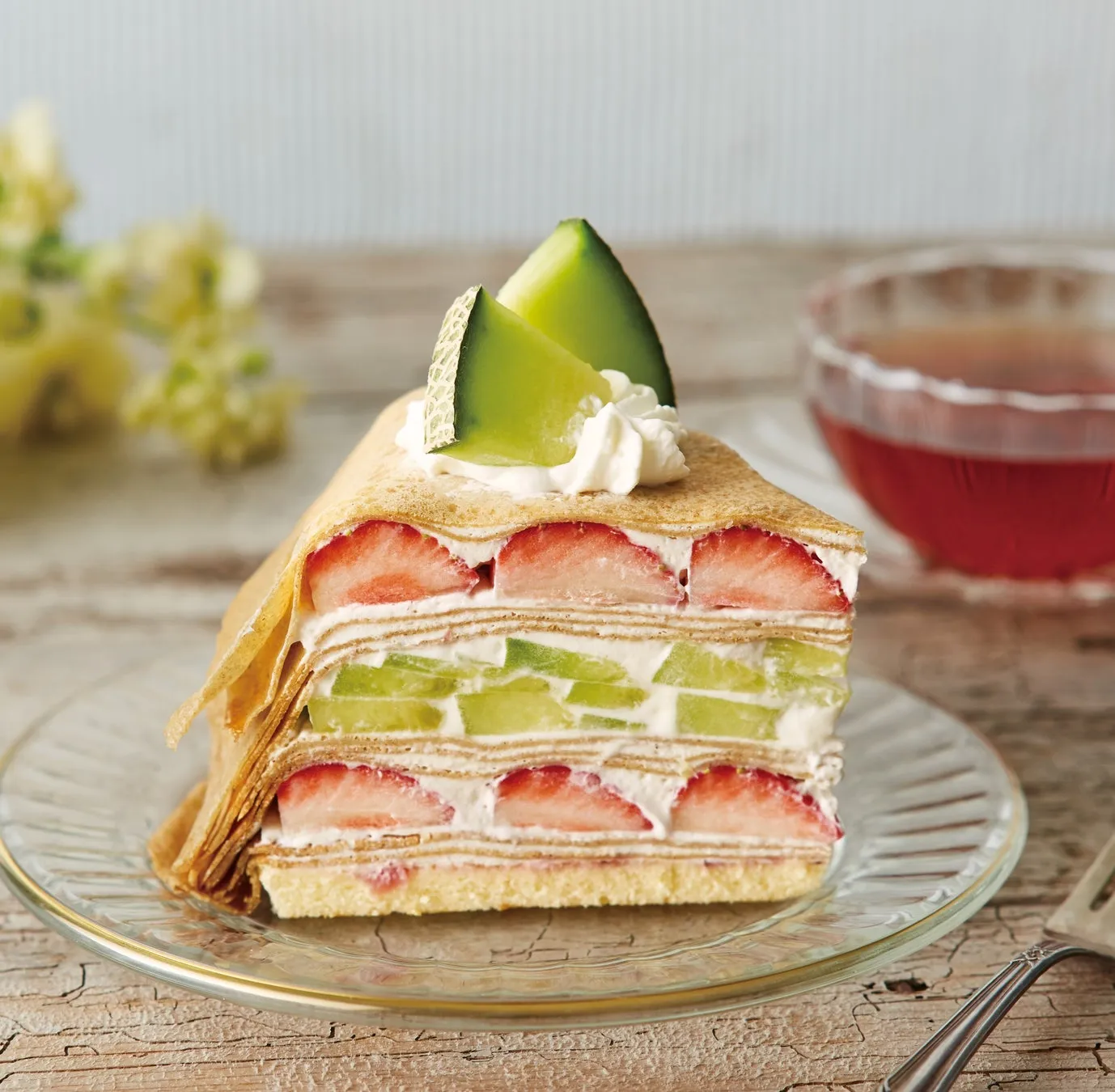 メロンとイチゴがトッピングされたクレープケーキがガラスの皿にのっている写真です。背景には、飲み物が入ったグラスと、木製のテーブルがあります。