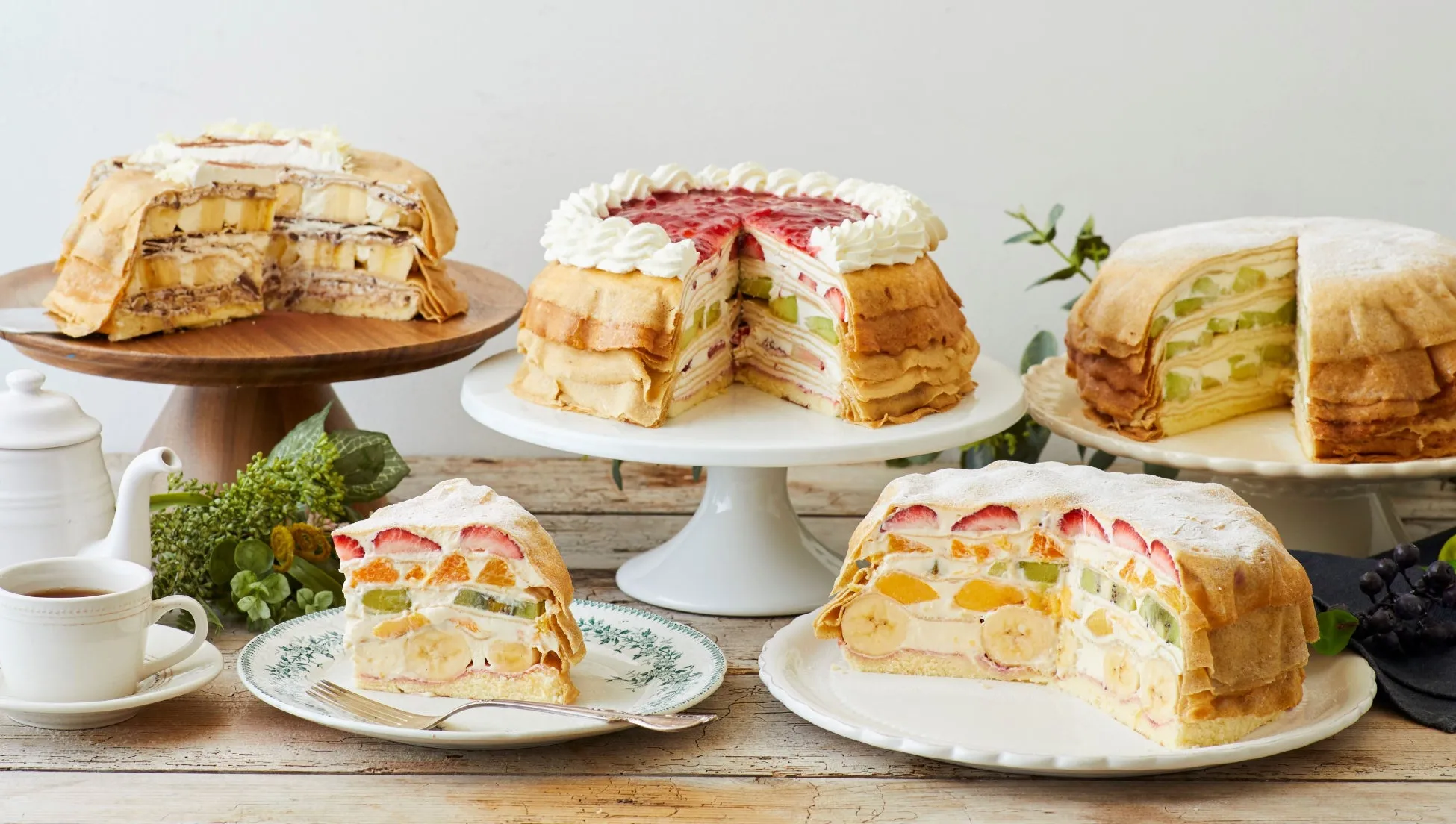 様々なフルーツがトッピングされたクレープケーキが複数並べられた写真です。カットされたケーキは断面が見えており、層になったクリームやフルーツが確認できます。ケーキはそれぞれ異なる台座に置かれており、一部は皿に盛り付けられています。テーブルの上には、ティーセットやフォーク、ブドウなどの装飾も配置されています。