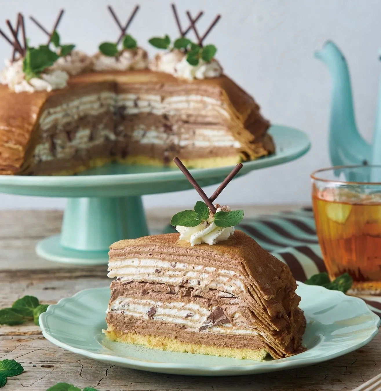チョコレートクレープケーキが皿に載っており、ケーキの切れ端が皿に添えられています。ケーキの周りにミントの葉とチョコレートのスティックが飾られています。ケーキの後ろには、ケーキ全体が載った台座があり、背景には水色のティーポットと飲み物が入ったグラスがあります。