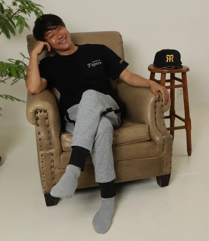椅子に座って笑顔を見せる男性。黒いTシャツに灰色のスウェットパンツ、野球帽を被っている。
