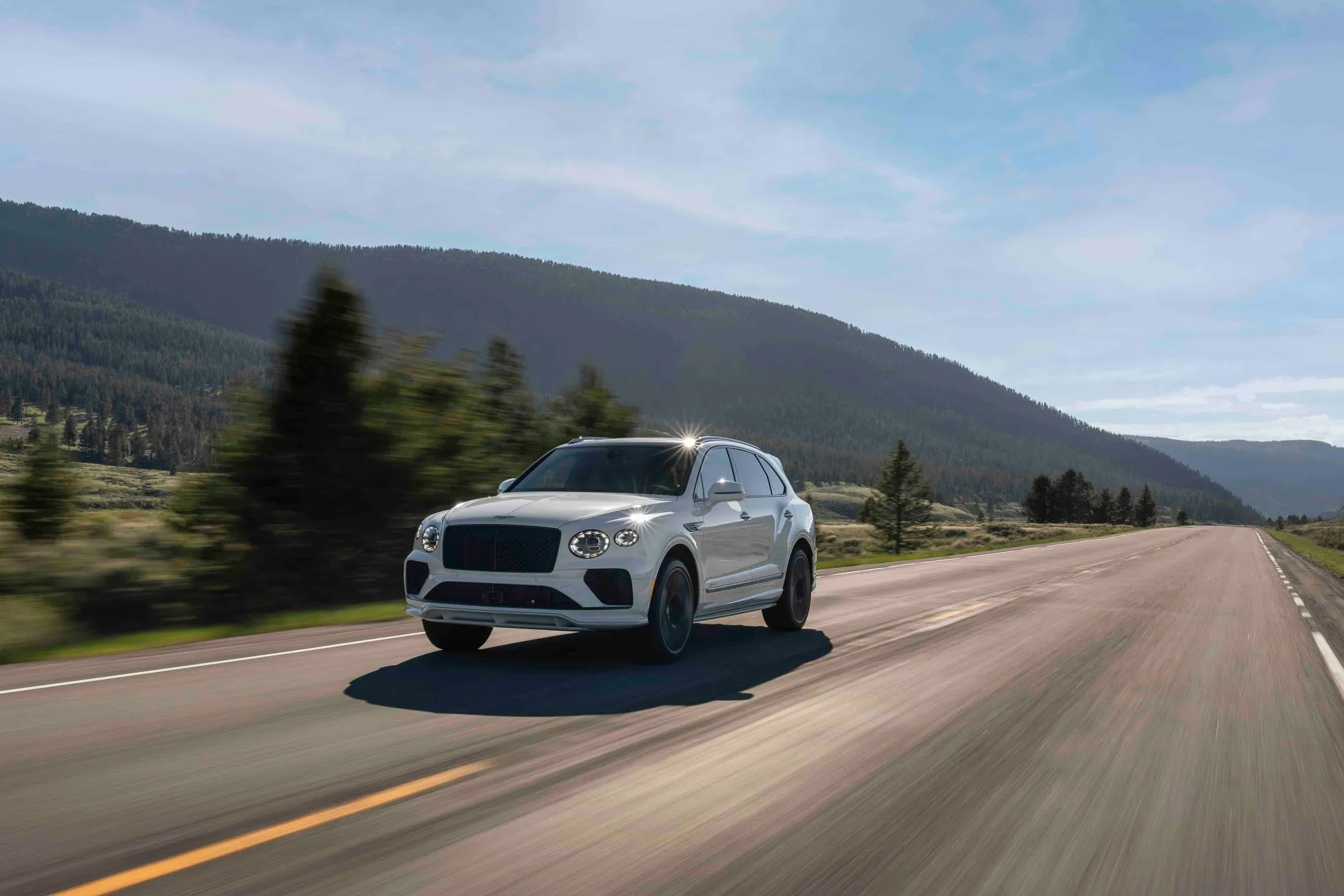白いBentley Bentaygaが、山と空を背景に、道路を走行している様子。