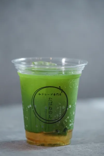 緑色の飲み物が入った透明な容器の写真です。下部には琥珀色の液体があり、上部には緑色の液体が注がれています。容器にはロゴマークと日本語の文字が印刷されています。