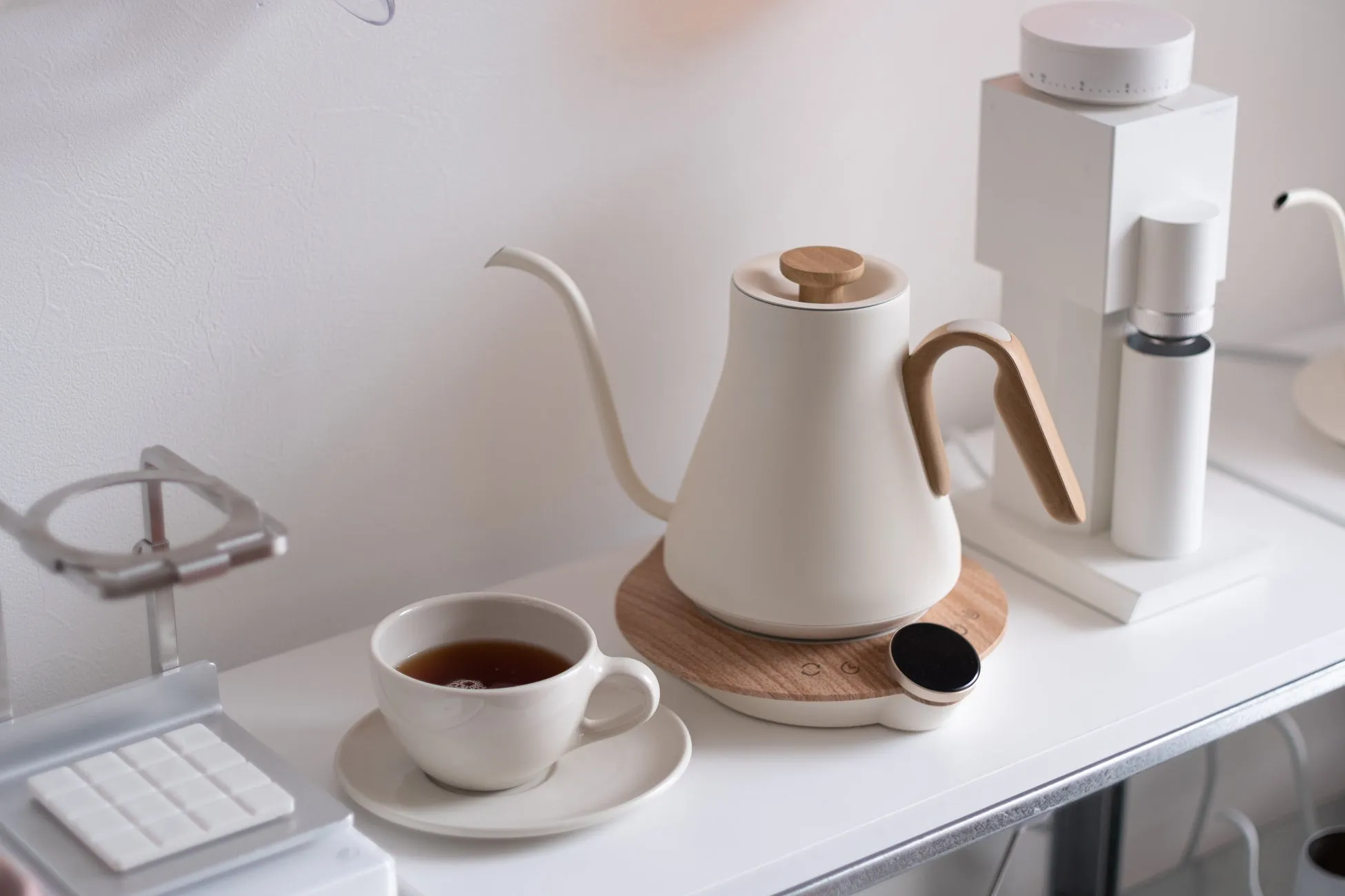 白い壁の前に置かれた、コーヒーポット、コーヒーミル、カップとソーサーに入ったお茶の静物写真。