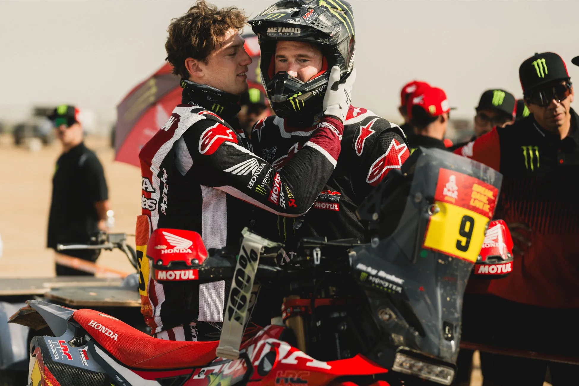 2人の男性がオートバイのレースの準備をしている様子。一人はヘルメットをかぶり、もう一人がヘルメットに手を添えている。バイクには「HONDA」や「MOTUL」のロゴが見える。