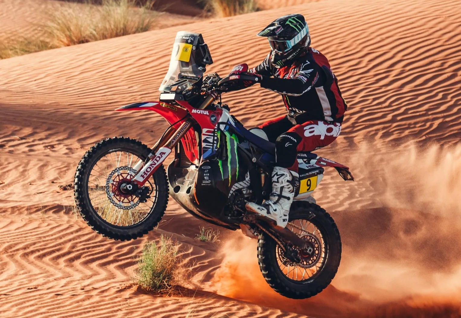 砂漠でバイクに乗っているレーサーの画像。バイクは空中にあり、ライダーはヘルメットをかぶっています。