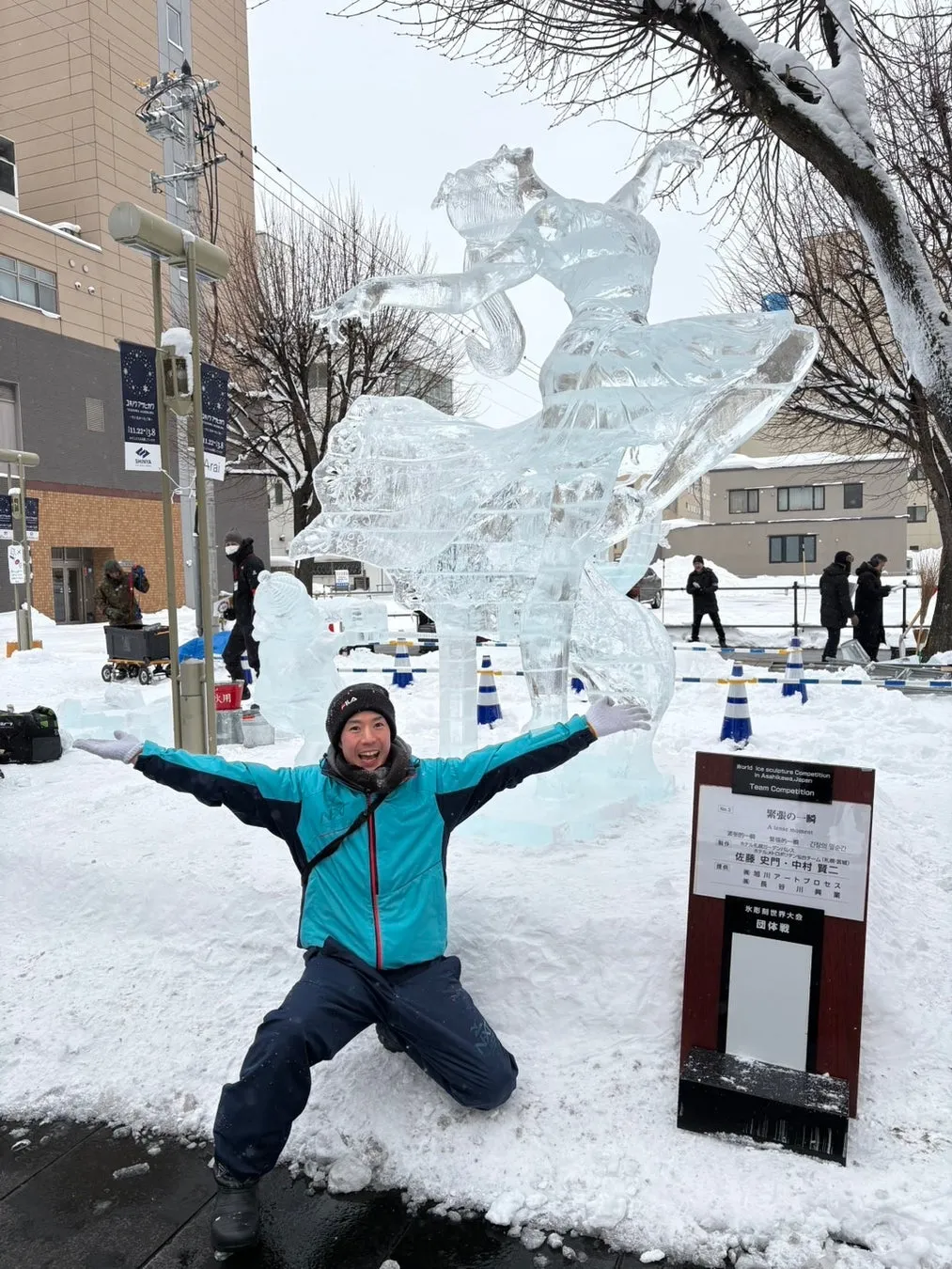 雪の中で、氷の彫刻の前にポーズをとる男。氷の彫刻はバレリーナを表現しています。男は青いジャケットを着ており、笑顔で両手を広げています。