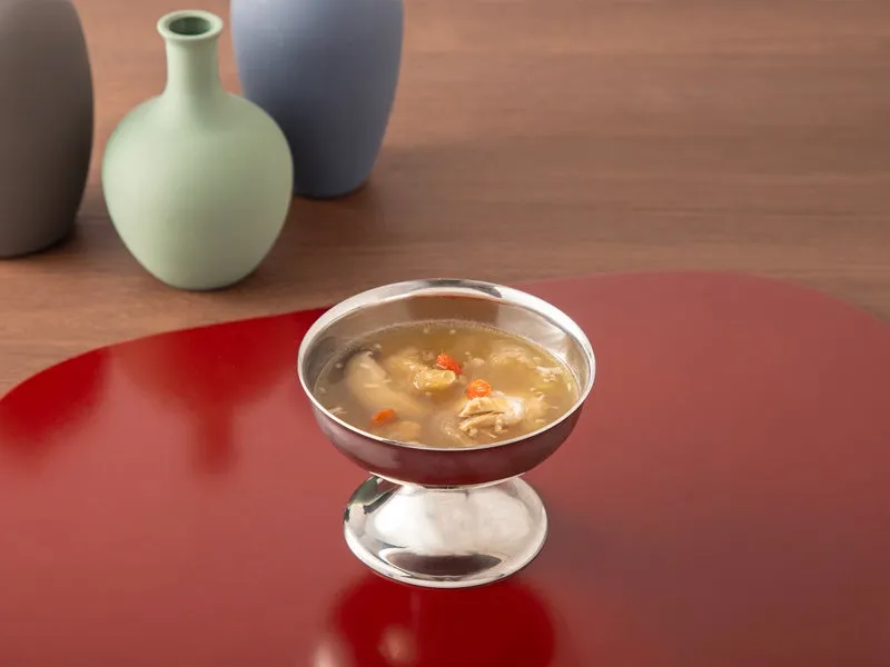 銀色の器に入ったスープのクローズアップ。背景には、3つの花瓶と赤いテーブルマットがある。