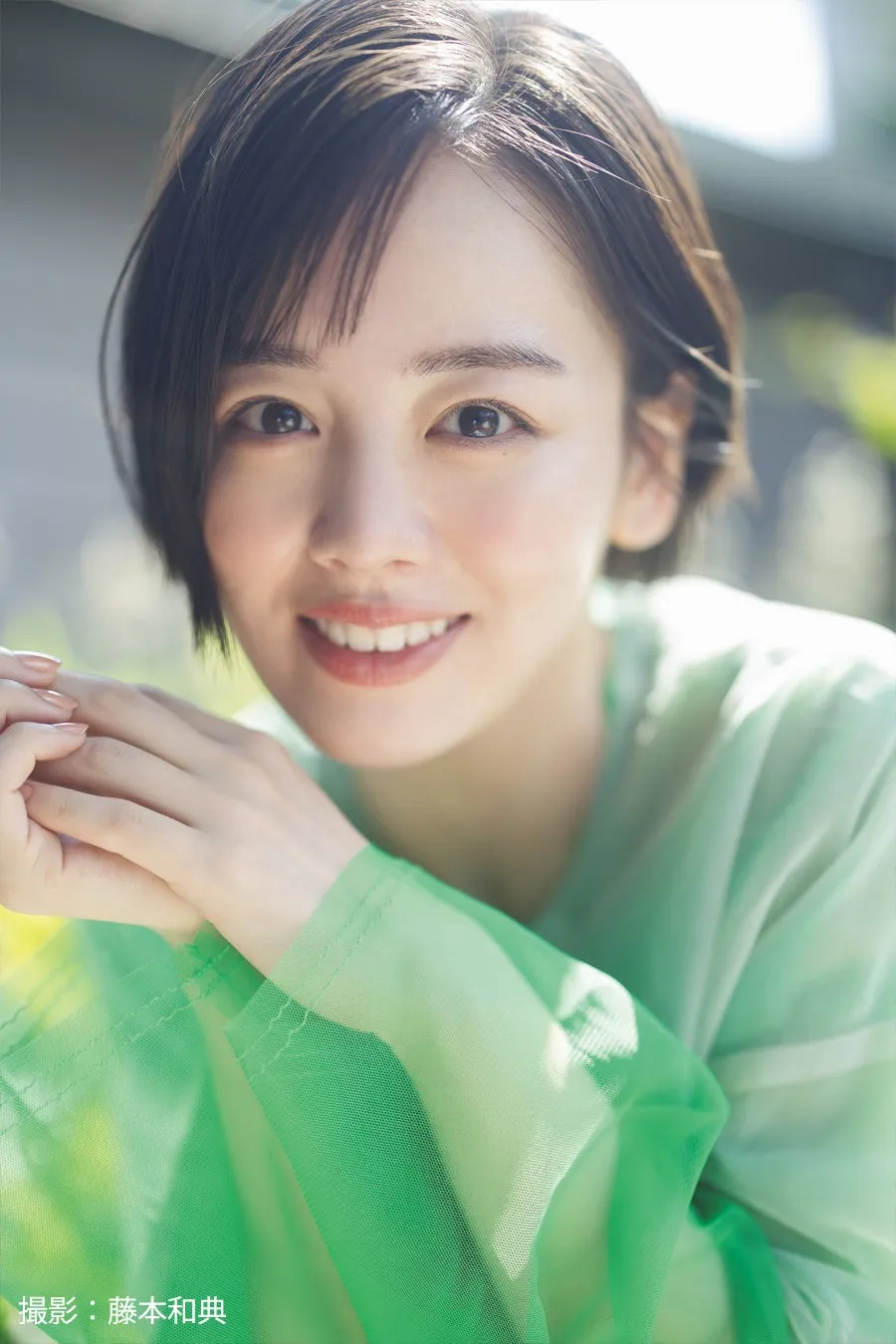 笑顔が印象的な女性のポートレート。薄いグリーンの服を着て、カメラ目線で微笑んでいる。背景にはぼやけた緑の木々が見える。