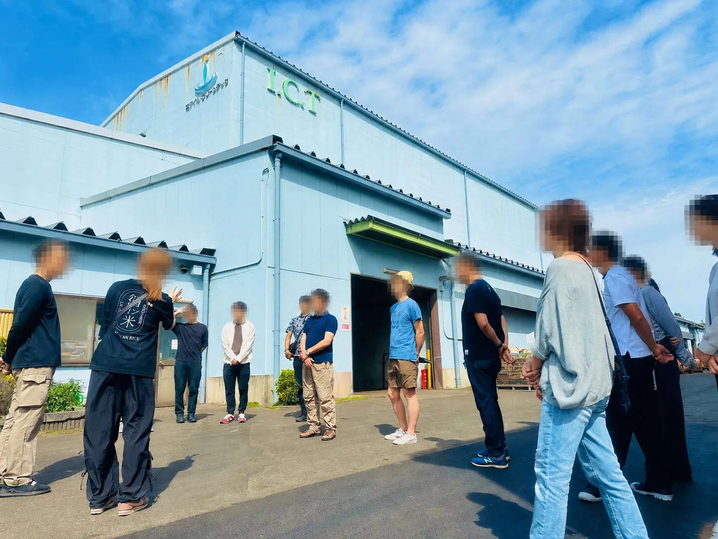 青い建物と人々のグループが屋外に立っている写真です。建物には「I.C.T」と書かれています。人々はカジュアルな服装をしており、建物を眺めているようです。