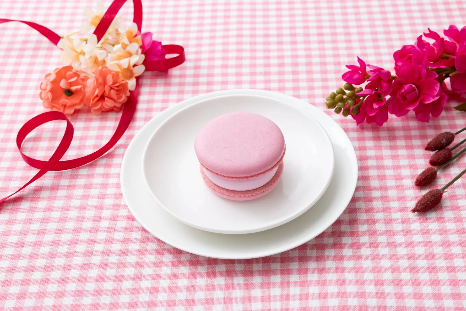 ピンクのマカロンが白い皿に置かれ、赤いリボンと花が添えられた、ピンクと白のチェック柄のテーブルクロスの上に置かれた写真