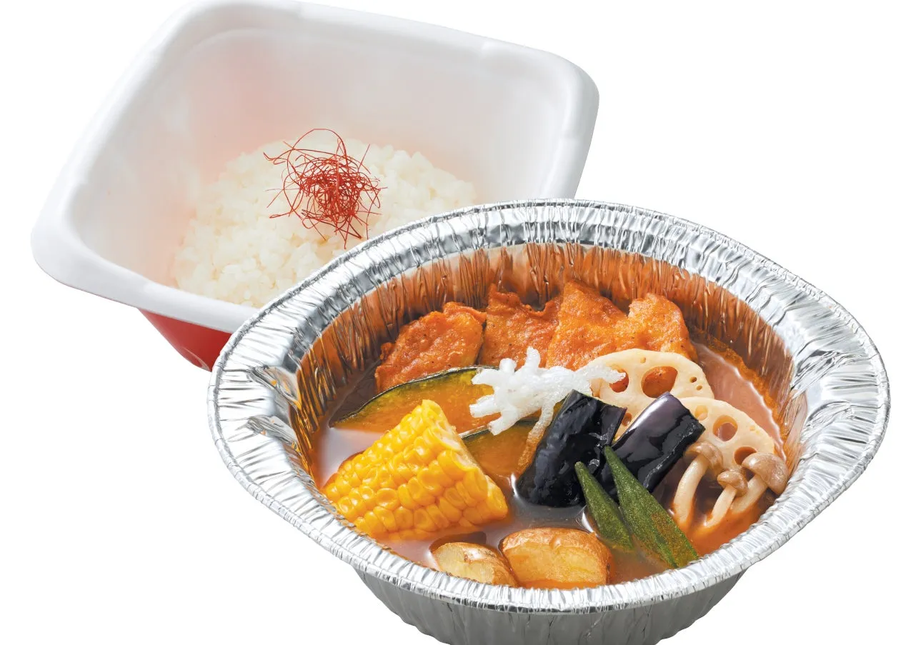 アルミホイルの容器に入ったスープカレーと、白いご飯のセットの画像。スープカレーには、揚げ物、野菜、きのこなどが入っている。
