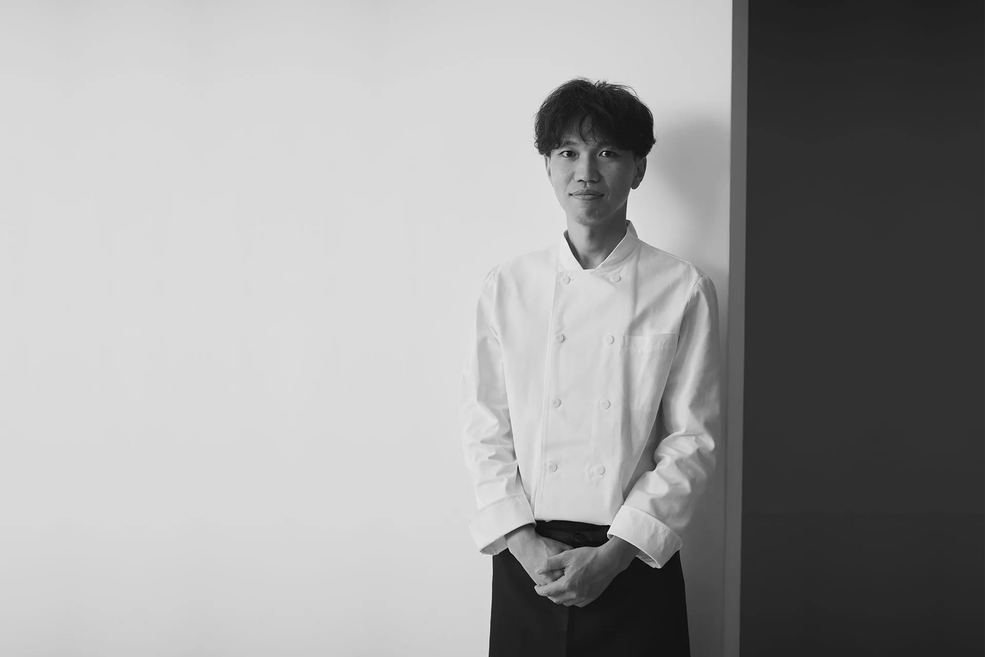 シェフの男性が壁に寄りかかって立っている白黒写真です。彼は白いシェフのコートと黒いエプロンを着ています。彼はカメラを見ています。
