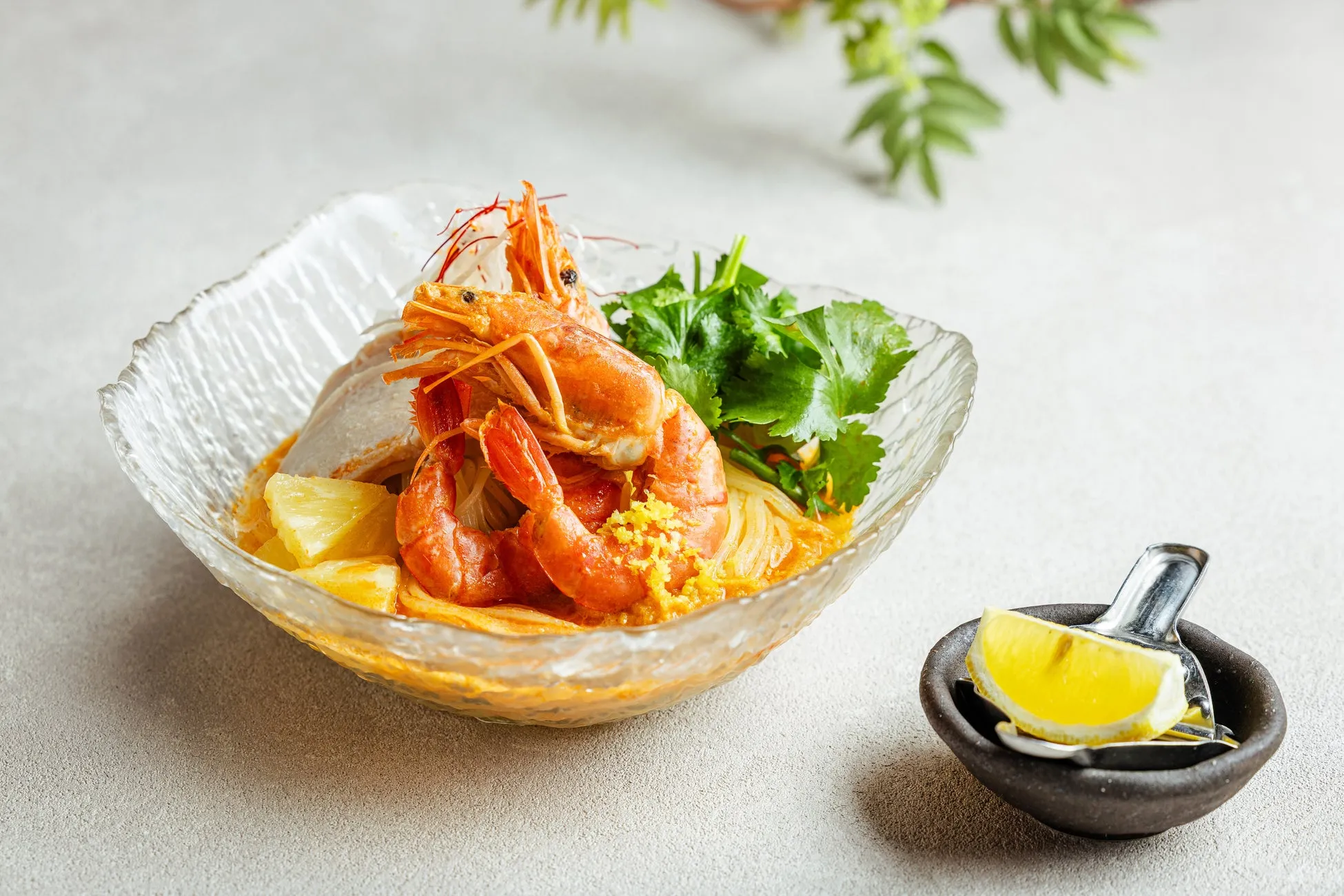 エビとパイナップル、パクチーが乗った麺料理。ガラスの器に入っており、横にはレモンが添えられている。