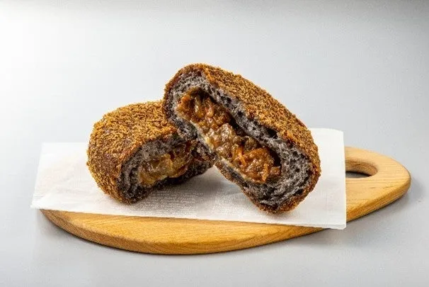 揚げパンが半分にカットされ、中身が見えるように配置されています。パンは茶色く揚げられ、中には茶色い具材が詰まっています。パンは木製のまな板の上に白い紙の上に置かれています。