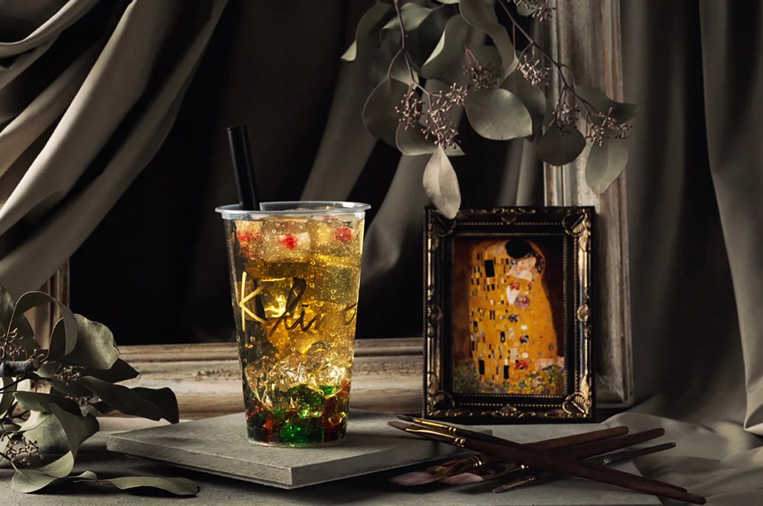 透明なプラスチックカップに入った、タピオカと氷が入った飲み物の写真。背景には絵画とブラシ、装飾的な葉が配置されている。