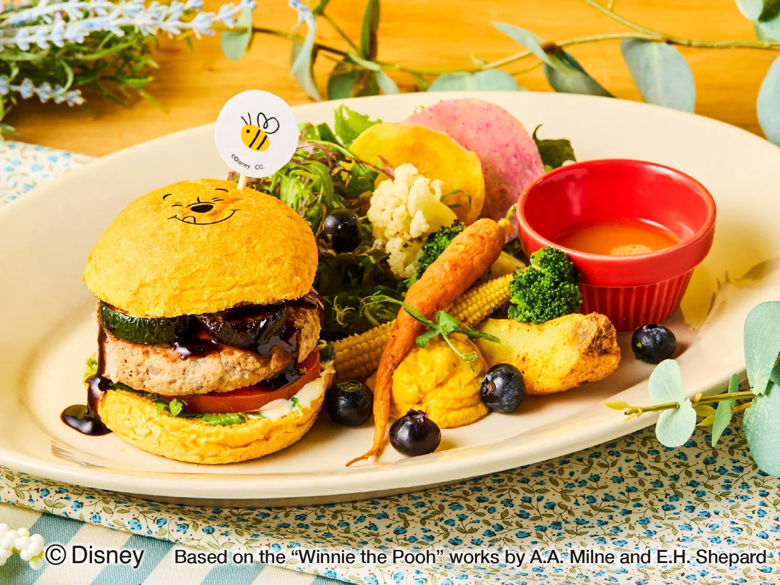 くまのプーさんの顔が描かれたハンバーガーと野菜の盛り合わせ。プレートには、ハンバーガー、野菜、ソースが添えられており、可愛らしい雰囲気で食欲をそそる。