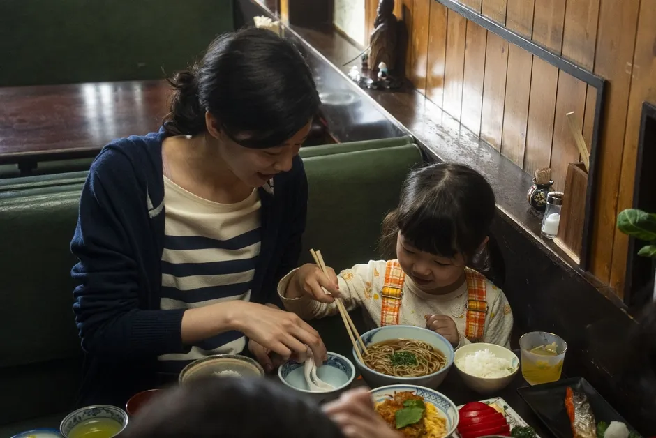 母親と子供が食事をしている様子。子供は箸を使って麺類を食べようとしており、母親はそれを見守っている。食事には麺類、ご飯、おかずなどが含まれている。
