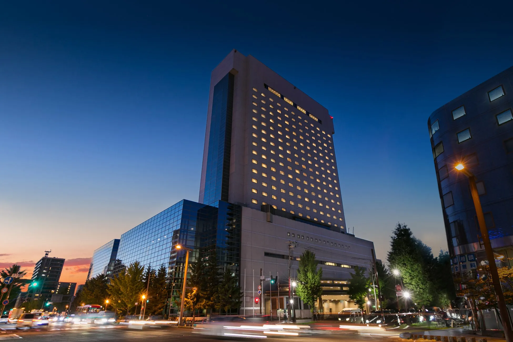 夜の街並み。高層ビルがそびえ立ち、街灯が周囲を照らしている。空は夕焼けと夜空が混ざり合い、美しいコントラストを生み出している。