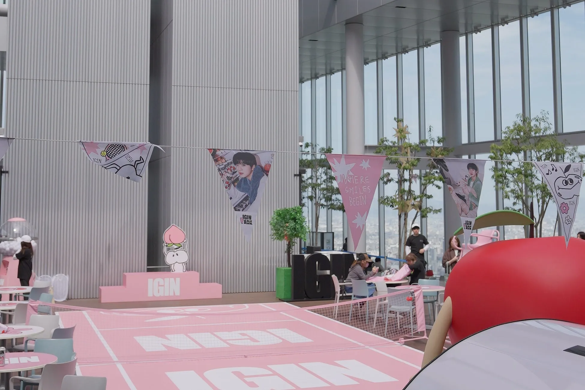 ピンク色のコートのようなエリアに、IGIN PARKのイベントスペースが設置されています。旗には人物の写真やIGINのロゴが描かれ、イベントのテーマを示唆しています。背景には高層ビルや木々が見え、屋外でのイベントであることがわかります。